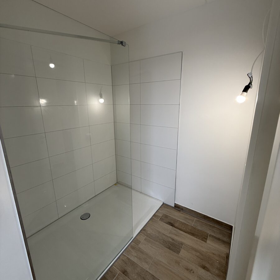 Moderne Dusche mit Glaswand, weißen Fliesen und stilvollem Holzoptik-Boden. Minimalistisch und elegant.
