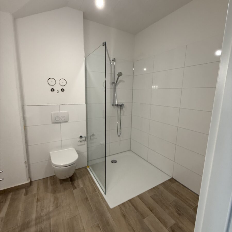Modernes Badezimmer mit eleganter Glasdusche, hölzernem Boden und minimalistischer Beleuchtung.